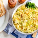 Eiersalat mit Sellerie in einer Schüssel, umgeben von Brot und Gewürzstreuern, ideal für eine gesunde Mahlzeit.